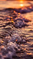Ocean waves with water droplets and bokeh lights during golden hour sunset creating warm orange and purple reflections on sea surface.