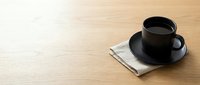 Black coffee cup and saucer on wooden table with napkin, minimalist workspace setup for cafes, offices, and lifestyle design projects.