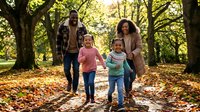 Two parents and two young children walking together through an autumn park surrounded by tall trees and fallen leaves, enjoying quality family time outdoors.