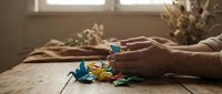 Elderly hands holding colorful paper origami cranes on wooden table with warm natural lighting and dried flowers in rustic home interior setting.