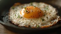 Crispy fried egg with bright orange yolk, seasoned with fresh herbs and black pepper in cast iron pan, macro food photography with soft lighting.