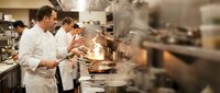Professional chefs cooking in busy commercial kitchen with flames and steam during dinner service preparation.