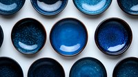 Ceramic bowls in shades of deep blue and turquoise arranged in rows on white background, showing handcrafted texture and glazed finish for modern table setting.