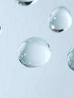 Clear water drops floating in air against light background, macro photography showing surface tension and liquid refraction with soft focus effect.