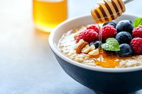 Creamy oatmeal topped with fresh raspberries, blueberries, sliced almonds and honey drizzle in dark bowl. Healthy breakfast with golden honey jar in background.
