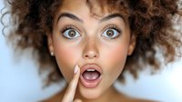 Young African American woman with curly afro hair expressing surprise with wide eyes and open mouth, touching face with finger against light background.