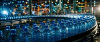 Rows of blue plastic bottles on industrial conveyor belt in manufacturing facility with blurred factory background and lighting.