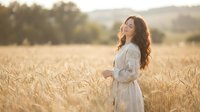 Young woman with long curly hair in white dress standing peacefully in golden wheat field during sunset, enjoying nature and freedom in countryside.