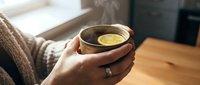 Person holding warm cup of tea or coffee with steam rising, wearing cozy sweater and ring, sitting at wooden table by window for relaxation and comfort.