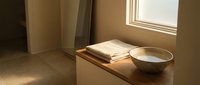 Minimalist interior with wooden shelf holding stacked papers and ceramic bowl, sunlight streaming through window creating warm shadows on beige walls.