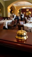 Golden service bell on wooden counter in upscale restaurant with blurred dining room and waitstaff in background for hospitality and customer service concepts.