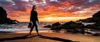 Man in wetsuit standing on beach at dramatic sunset with rocky coastline, ready for surfing or water sports adventure.
