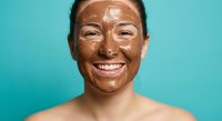 Young Asian woman with chocolate face mask smiling against turquoise background. Natural skincare beauty treatment for healthy glowing skin and wellness routine.