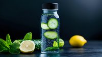 Fresh cucumber and mint infused water in glass bottle with lemon on dark background, healthy detox drink with bubbles and sliced ingredients.