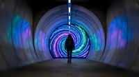 Person silhouette walking through futuristic tunnel with colorful neon light rings creating spiral motion blur effect in modern underground passage.