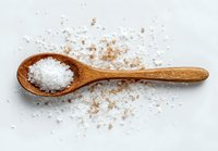 Wooden spoon with sea salt crystals and scattered pink Himalayan salt on white background for culinary and wellness concepts.