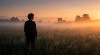 Young man silhouette standing in misty field at golden sunrise with warm atmospheric light creating peaceful morning landscape scene.