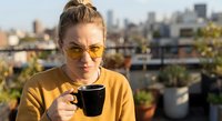 Young blonde woman in yellow sweater enjoying morning coffee on urban rooftop terrace with city skyline background during golden hour sunrise.
