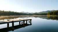 Wooden dock extending into calm lake with forested mountains and clear sky reflected in still water, suitable for travel, nature, and outdoor recreation marketing.