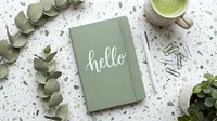 Sage green notebook with hello lettering on terrazzo surface, surrounded by eucalyptus, matcha latte, pen and paper clips.