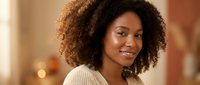 Young African American woman with curly hair smiling warmly in cozy sweater at home. Natural portrait for lifestyle and wellness content.