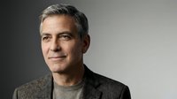 Middle-aged man with gray hair and facial stubble wearing casual brown blazer over gray shirt, studio portrait against neutral background.