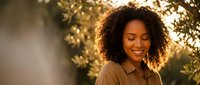 Young African American woman with curly hair smiling peacefully outdoors in warm golden sunlight among green foliage creating serene natural portrait moment.