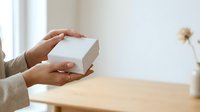 Woman with medium skin tone holding white gift box with both hands at wooden table, suitable for e-commerce, retail, and product packaging presentations.