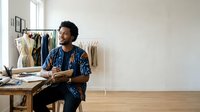 Young male fashion designer with curly dark hair and medium brown skin tone, wearing blue patterned shirt, sitting at wooden desk holding fabric swatches in bright studio workspace with clothing rack and design materials.