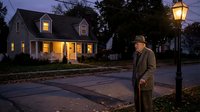 Elderly Caucasian man in coat standing on suburban street at dusk with warm house lights and vintage street lamp creating cozy neighborhood atmosphere.