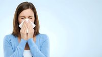 Young woman with brown hair and fair complexion wearing light blue sweater blowing nose with tissue on white background. Healthcare and cold season.