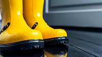 Bright yellow rubber rain boots with water drops on black surface, close-up view showing glossy wet texture and reflection. Essential protective footwear for rainy weather.