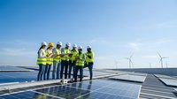 Diverse team of engineers and technicians in safety vests inspecting solar panel installation at renewable energy facility with wind turbines.
