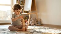 Young toddler boy with curly hair playing with wooden building blocks in bright nursery room with natural sunlight and cozy home interior.