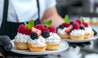 Fresh berry tartlets topped with whipped cream, raspberries, blackberries and mint leaves on white plate. Baker holding dessert platter in restaurant kitchen.