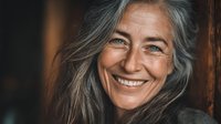 Close-up portrait of mature woman with long gray hair and bright smile showing natural beauty and confidence against dark background.