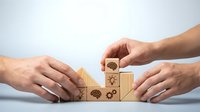 Hands arranging wooden blocks with brain, gear, and lightbulb icons into staircase steps, symbolizing teamwork, creativity, and business growth.