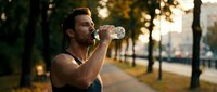 Athletic man drinking water from bottle during outdoor workout session in urban park setting with golden hour lighting for fitness and hydration concepts.