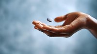 Close-up of human hand with small floating pill or tablet suspended in air against blurred blue background, demonstrating gravity defying pharmaceutical concept.