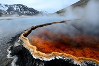 Vibrant orange and red geothermal hot spring with mineral deposits and rising steam surrounded by snow-capped mountains, dramatic volcanic landscape in winter setting.