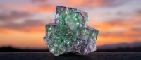 Fluorite crystal mineral specimen with green and purple colors against dramatic sunset sky background for geology education and gemstone collection displays.