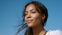 Young mixed race woman with glowing skin smiling against bright blue sky background, wearing white top, natural makeup and windswept hair for beauty and lifestyle content.
