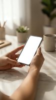 Hands holding smartphone with blank white screen mockup on marble desk in bright modern office workspace with coffee cup and plants in background.