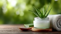 Fresh aloe vera plant in glass jar with natural cream, wooden spoon and towel on rustic table against blurred green background for spa and skincare.