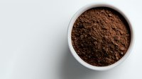 Ground dark cocoa powder in white ceramic bowl on light background, overhead view showing rich brown color and fine texture for food and beverage designs.