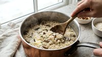 Person stirring oatmeal mixture in metal pot during breakfast preparation near window with natural light.