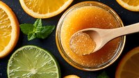 Fresh citrus jam with orange slices, lime and mint leaves on dark background. Wooden spoon shows crystallized texture of homemade marmalade preserve.