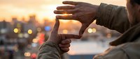 Male photographer framing sunset cityscape with hands creating viewfinder gesture during golden hour with bokeh lights and urban skyline background.