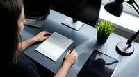 Young woman with long dark hair, East Asian ethnicity, working at modern desk with laptop, notebook and pen, taking notes or writing, professional office environment with desk lamp and potted plant, overhead view.