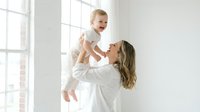 Young Caucasian mother in white shirt lifting laughing baby in bright minimal room, sharing joyful bonding moment full of love and happiness.